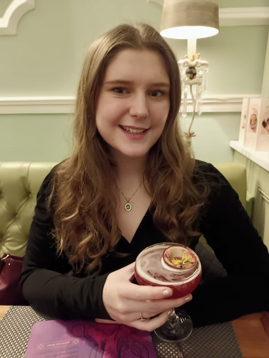  A close-up image of Molly sitting at a table in a restaurant. She is smiling and holding a passion-fruit cocktail. She has long, light hair and is wearing a dark top.