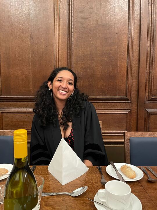 A picture of Ananya sitting in at a table the Great Hall at a formal. She has curly black hair and is smiling.