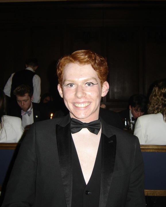 A headshot of Harry with a formal dinner in the background. He is wearing a black dinner jacket, waistcoat, and bowtie, he has ginger hair and has black eyeliner on.