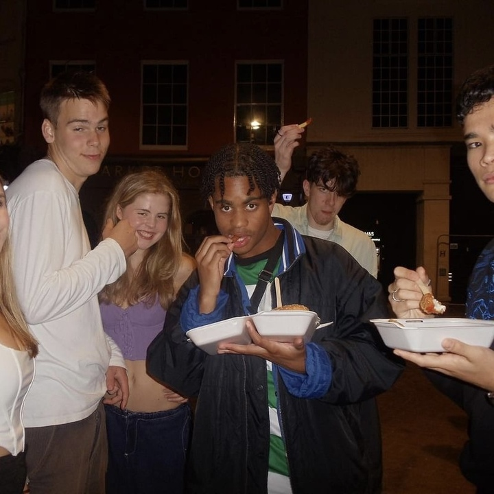 Picture of Enyioma outside in a black Nike jacket and football shirt with some of Enyioma's friends in the background. He is midway through eating a burger and chips and is looking into the camera.
