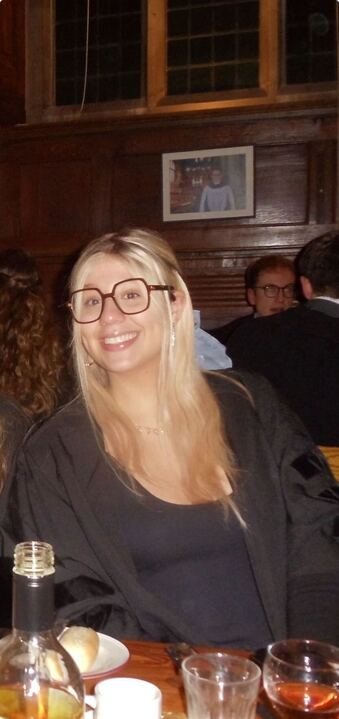 A photo of Erin sat down at a formal. She has blonde hair, big glasses, and a black dress with a Clare gown on!
