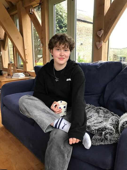 A photo of Luca sat on a sofa with a mug. They have short curly brown hair and are wearing a hoodie and grey cords.