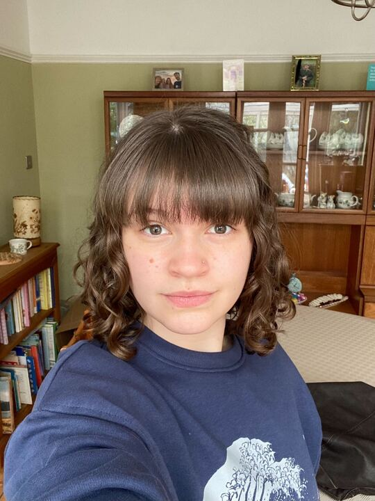 A selfie of Francesca wearing a blue sweatshirt with an image of a wave on it. She has shoulder-length brown curly hair, a fringe, and a nose-stud. In the background is a bookcase and a wooden dresser containing crockery.