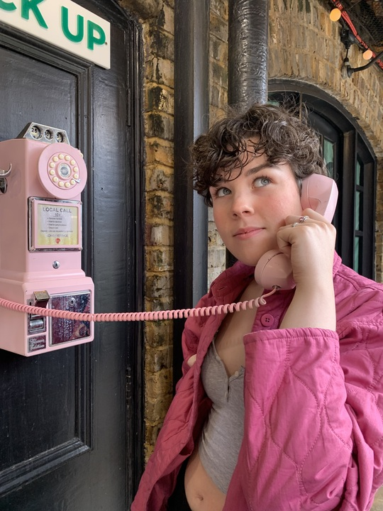 A picture of Niamh. She has short brown hair, is wearing a grey top and a pink jacket and holding a pink phone.