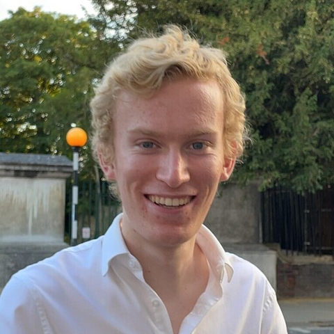 A headshot of Thomas with a street and greenery in the background. He has short blond hair and a white shirt, and is smiling.