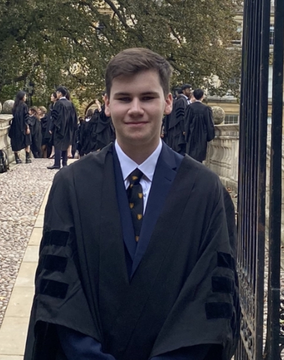 A mid-shot of Max standing in the gate of Clare Bridge. He has short brown hair and wears a gown. Other post-matriculation students can be seen in the background.