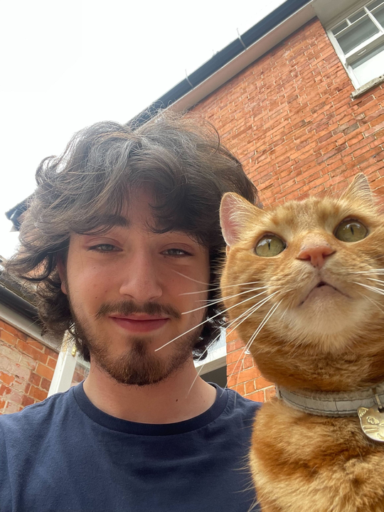 A selfie of Luca outside a house with a ginger tabby cat. Luca has medium-length brown hair, facial hair, and a blue t-shirt.