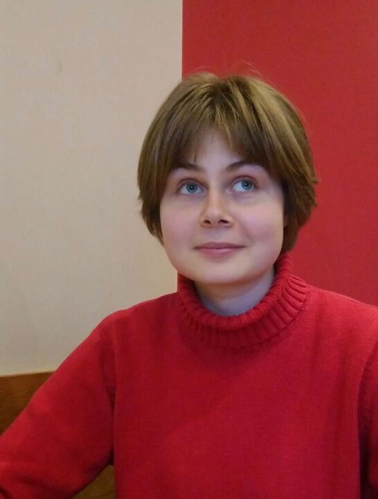 Meryn is wearing a red turtleneck jumper against a red and white background. She has short light brown hair and is looking up, away from the camera.