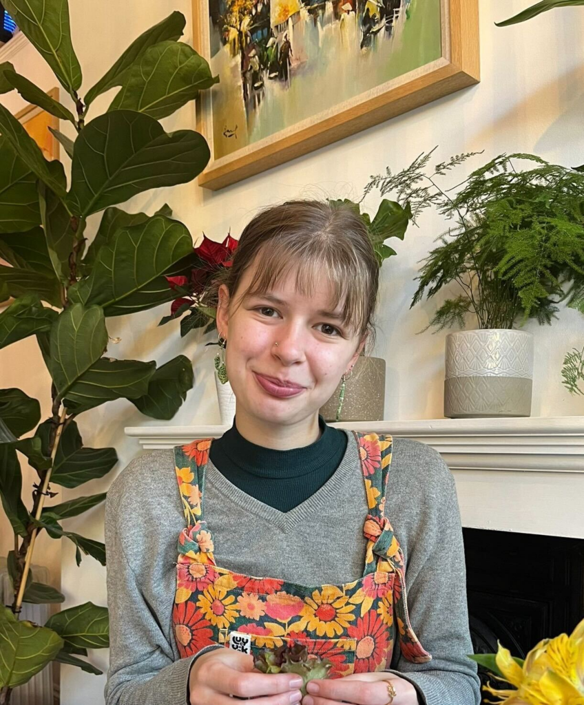 A mid shot of Cat with lots of houseplants in the background. She has a fringe, colourful dungarees, and she is smiling at the camera.
