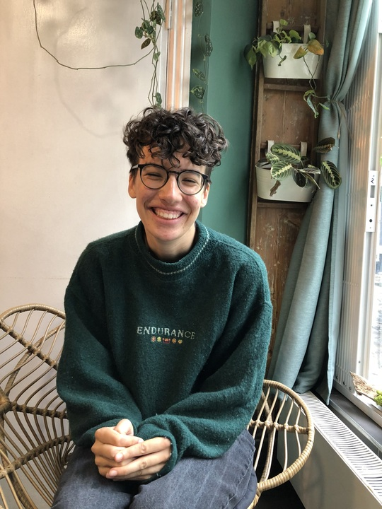 A mid-shot of Mary with a background of plants on a wall. They have short curly brown hair, a green jumper, brown glasses and they are smiling at the camera.