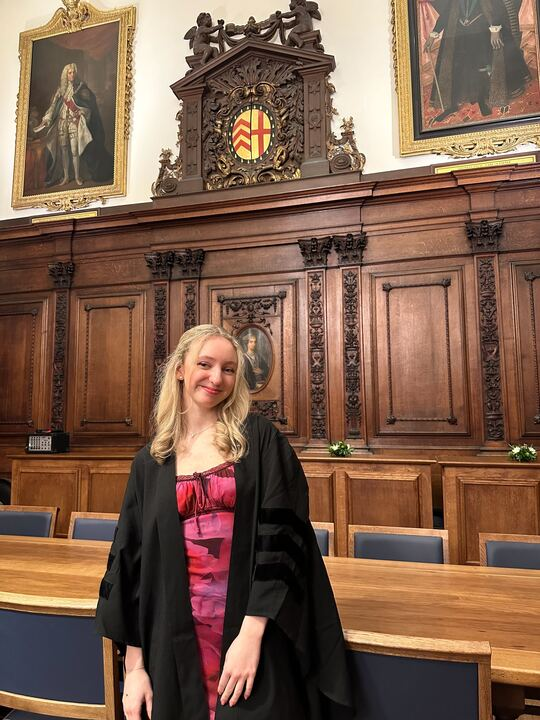 A mid-shot of Isabella standing in front of the head table in the Great Hall. She is smiling and has medium-length blonde hair and a pink dress. The façade and paintings of the Great Hall's back wall are visible in the background.
